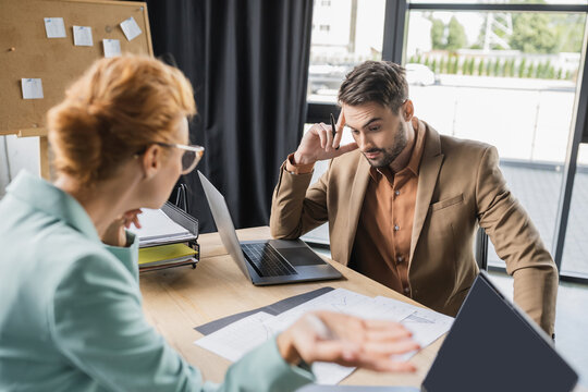 discouraged manager looking at papers near blurred businesswoman gesturing during conflict in office.