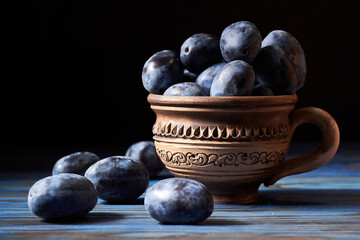 Fresh black plums in a clay rustic mug against dark blue background