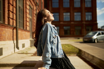 Fototapeta Side view outdoor image of gorgeous student girl with closed eyes in denim outfit leaning on edging at city streets taking sun bathing with face turned to sunlight, enjoying warm day and freedom obraz