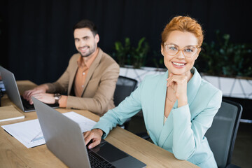 joyful businesswoman smiling at camera near laptop and blurred colleague in office.