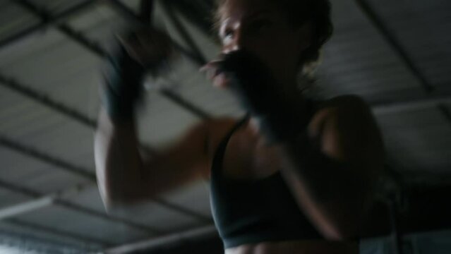 Woman boxer training at gym in the dark. Female fighter with curly hair and wearing black sportswear practicing punches and kicks in the ring