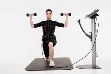 Fit girl does lunges with dumbbells physical exercises in EMS suit that uses electrical impulses to...