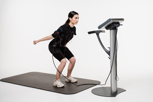 Fitness girl in EMS suit is doing workout sport training that uses electrical impulses to stimulate muscles on white background. Sport training in electrical muscle stimulation suit.