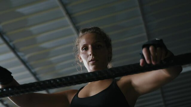 Female boxer leaning on ring ropes in gym looking at camera. Caucasian female muay thai fighter wearing black sportswear and boxing bandages takes a breath between fights