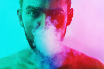 Fototapeta premium Handsome young man with wet skin in colorful light smoking cigarette or marijuana
