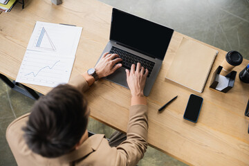 top view of brunette businessman typing on laptop with blank screen near documents and smartphone in office.