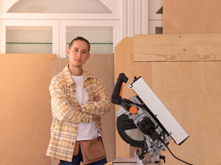 Young male carpenter with arms crossed standing in home carpentry shop against wood planks near...