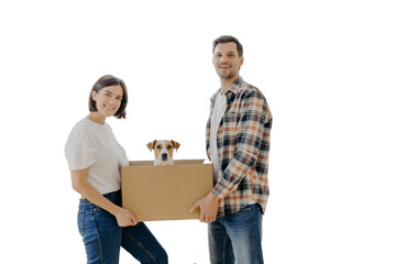 Fototapeta na wymiar Happy family couple hold carton box with small puppy, stand indoor against big window, glad to become homeowners, unpack things in own apartment, looks gladfully at camera. Family moving day