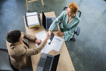 top view of successful partners shaking hands near business analytics and laptops in office.