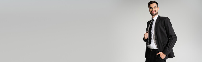 successful and happy businessman in black formal wear posing with hand in pocket isolated on grey, banner.