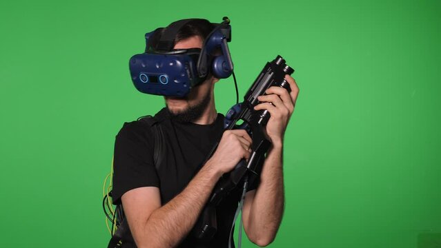 A man in a VR headset for video games with a glasses and a weapon on a green background, he takes aim from a machine gun. A man with equipment for playing in virtual reality in the Chroma key studio.