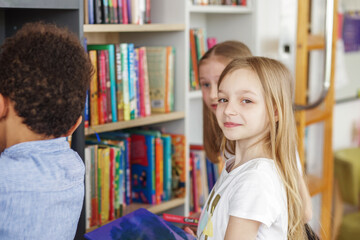 Three multiethnic children look for books near bookshelves and read together in school library.