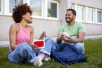 Two young students of different ethnicities are sitting on an outdoor lawn having a meal. The girl...