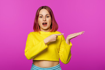 Happy smiling young woman with pink hair looking at camera and pointing with finger at copy space on pink background studio