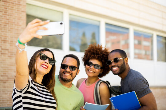 A group of young millennial students of different ethnicities are outdoors. The young people are taking a picture with the mobile phone with sunglasses on. Multiracial group photo concept.