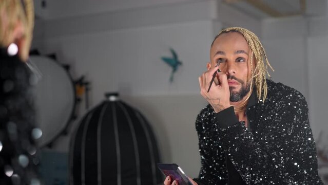 Gay man applying eyelashes looking mirror dressed black jacket with sequins . Brazilian black ethnicity homosexual guy with beard doing make up. Non-binary gender person. LGBT people gender identity