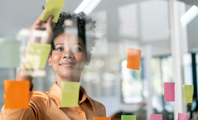 Young smiley attractive african businesswoman using sticky notes in glass wall to writing strategy...
