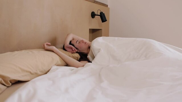 Side view of Young brunette woman sleeps peacefully on bed, wakes up, stretches and goes back to sleep in the morning. Doesn't want to wake up. Horizontal, hd video 4k resolution.