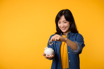 Happy young asian woman wearing yellow t-shirt denim shirt saving digital crypto currency to piggy bank isolated on yellow background. Digital money saving investment concept.