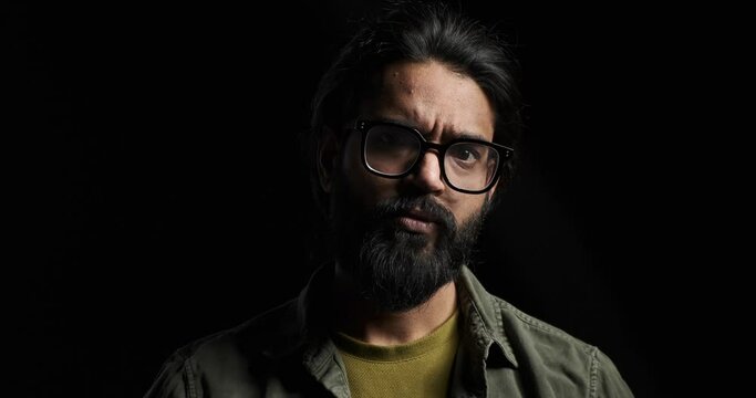 Slow motion shot of angry bearded young man with eyeglasses shaking head and staring seriously at camera against black background