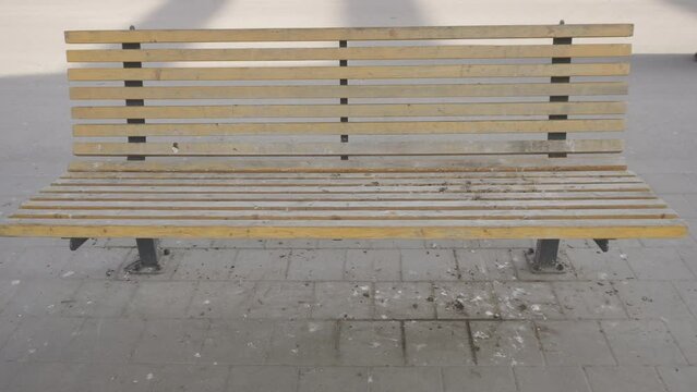Dirty empty wooden bench in city on tiled pavement, handheld shot. Bird droppings have soiled and rendered unusable city seat for resting and waiting. Unrecognizable woman passes by.