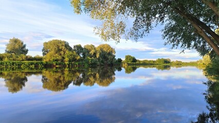 A beautiful cloudy sky in the evening over the river. The branches of trees and bushes lean over the water in which the trees from the opposite grassy bank are reflected, illuminated by the sun