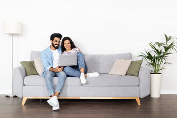 Loving eastern couple sitting on couch at home, using laptop