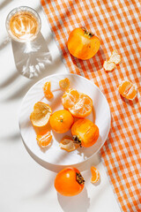 Creative composition made of fresh orange tangerin, persimmon fruits and a glass of drink on white background  with palm tree leaf shadow. Healthy food concept. Summer picnic idea.