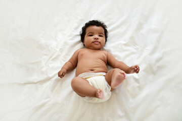 Glad cute african american little curly baby in diaper lies on white soft white bed, waking after sleep - obrazy, fototapety, plakaty