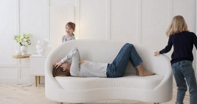 Mother with headache lying on sofa while mischievous daughters running around her and playing in living room