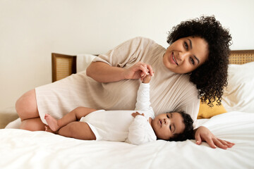 Smiling young african american female and little cute child lie on bed, relax together, play in bedroom