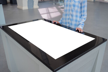 Woman in blue plaid shirt looking at blank interactive touchscreen white display of electronic table kiosk at exhibition or museum. White screen, mock up, copyspace, template, technology concept.