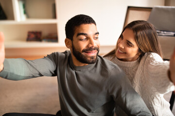 Glad millennial international husband and wife have fun, take selfie on phone together in living room interior