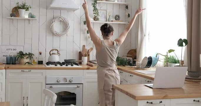 Carefree woman playing her favourite song using laptop and dancing while singing song at the kitchen