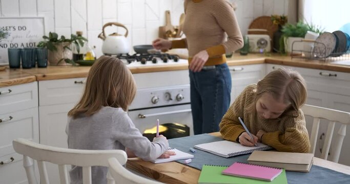 Smiling caucasian young mother cooking food and assisting daughters in solving mathematics problems in kitchen at home