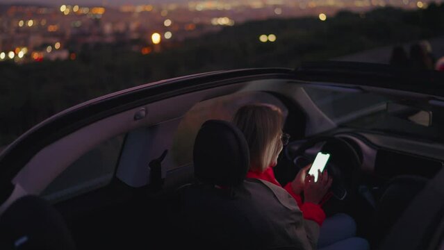 Smiling female influencer sits comfortably in convertible car,scrolling through an app on cellphone to share publication, communicate with social media network. Scenery night city view in background