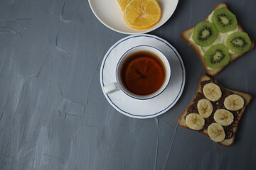 breakfast, tea with lemon and sandwiches, bread with chocolate, banana and kiwi slices on a gray background with space for text