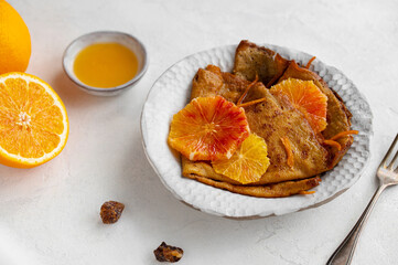 Traditional French Crêpes suzette pancakes with orange sauce on a serving dish on a light grey tale background decorated with citruses and bloody oranges.