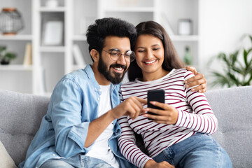 Relaxed multicultural couple sitting on sofa at home, using smartphone