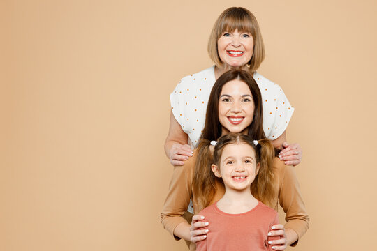 Happy smiling fun women wear casual clothes with child kid girl 6-7 years old. Granny mother daughter stand behind each other look camera isolated on plain beige background. Family parent day concept.