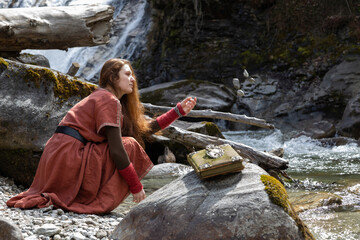 A red-haired young woman in an old dress sits on the bank of a mountain river of a rock and throws stones into the water