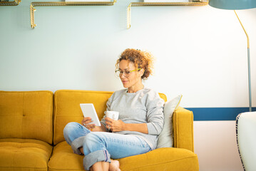 Smart casual woman middle age relaxing at home sitting on yellow sofa and using ereader to read an ebook enjoying indoor leisure activity alone. Sunday time lifestyle. People and technology online