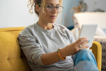 Attractive middle age woman portrait at home reading book on ereader modern device witting comfy on the yellow sofa. Female people in relax indoor leisure activity alone using technology smart tablet