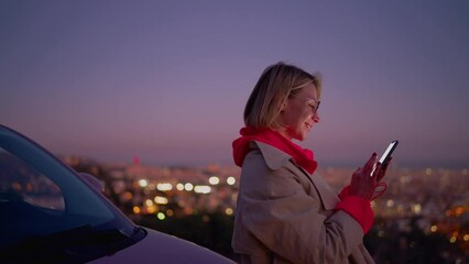 Blonde hair smiling female surfing 6G internet on smartphone while sitting in car at night time. Cheerful happy young woman making online payment while using cellular application. Modern technology - Powered by Adobe