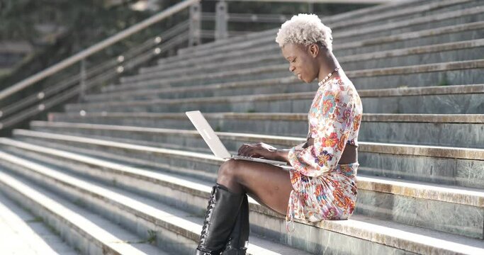 Positive black transgender person typing on laptop on stairs