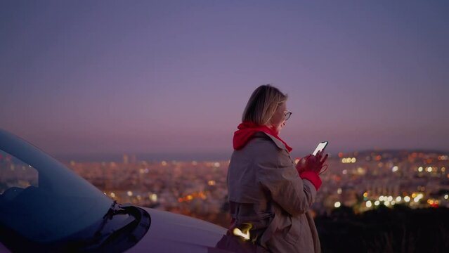 Smiling happy female browsing social media page on modern smartphone application with unlimited internet surfing. Carefree young woman enjoying online content at night city viewpoint near parked car