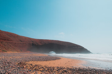 Fototapeta na wymiar Remaining red sandstone stone arch in Legzira Beach. Rugged coastline in Tiznit Province of Morocco, Africa. Atlantic Ocean waves. Travel background. Summer day. Empty shore. Lonely planet.