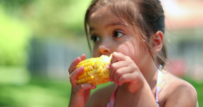 Cute little girl eating corn cob outside in nature. Small child girl eats healthy snack outdoors