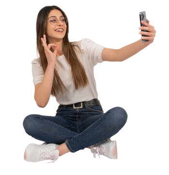 Woman doing selfie, full length portrait of young caucasian woman doing selfie. Sitting on the floor doing ok sign with hand. Using mobile phone. Transparent png image. Lifestyle concept idea.