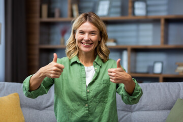 Portrait of a happy senior woman in a green dress sitting on the couch at home and smiling and showing super hands to the camera. - obrazy, fototapety, plakaty